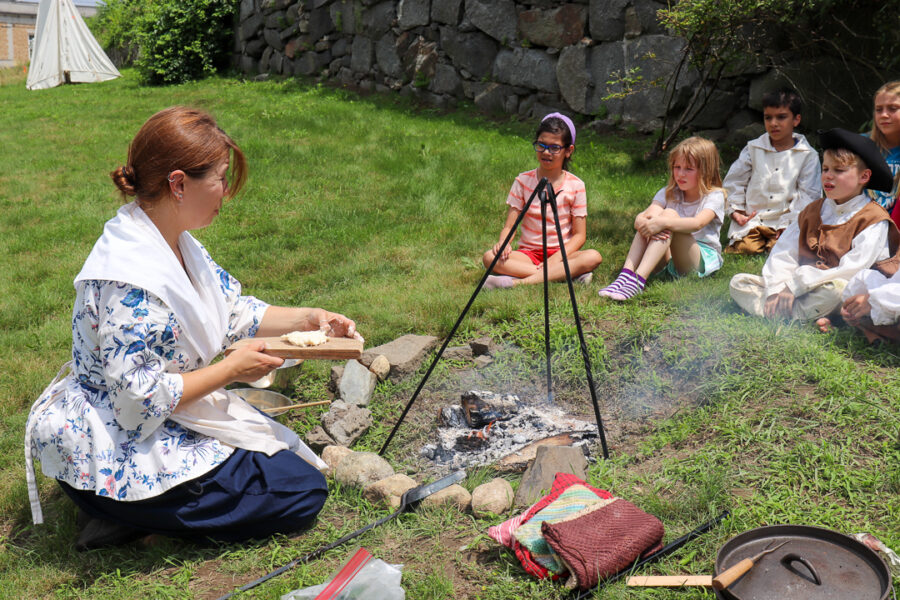 An adult dressed in 18th century clothing sits by an open camp fire and bakes flatbreads using the embers, a ring of children watch
