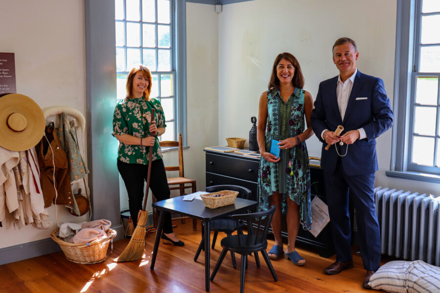 Three individuals stand in a room full of child friendly 18th century reproductions including a wall of dress up garments.