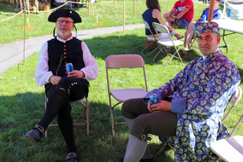 Two men in Revolutionary Era clothing sit and drink a beer